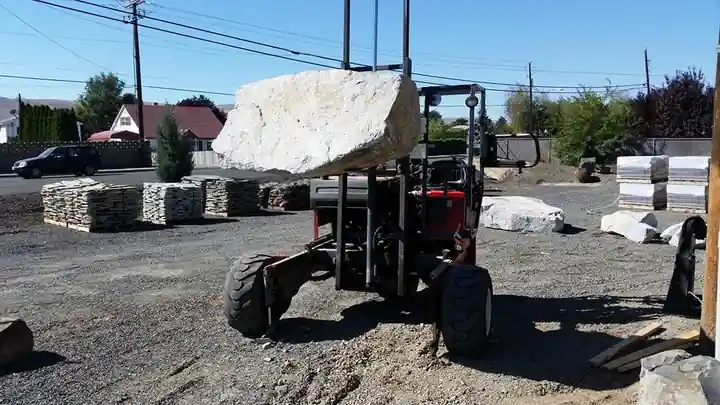 Truck-mounted forklift carrying a boulder across the gravel yard